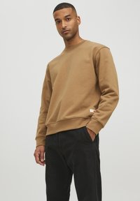 Brauner Sweatshirt mit rundem Ausschnitt, gerippten Bündchen und Saum. Verfügt über ein kleines Markenzeichen an der unteren Seite. Kombiniert mit schwarzen Hosen.