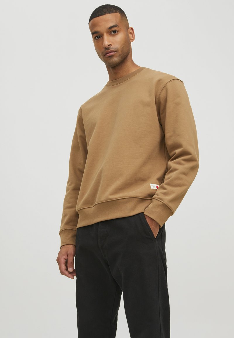 Brauner Sweatshirt mit rundem Ausschnitt, gerippten Bündchen und Saum. Verfügt über ein kleines Markenzeichen an der unteren Seite. Kombiniert mit schwarzen Hosen.