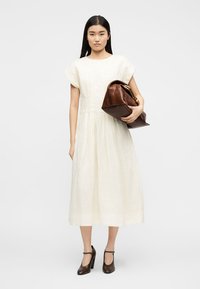 Femeie purtând o rochie midi albă și pantofi cu toc închiși, ținând sub braț o geantă mare de piele maro, stând în fața unui fundal simplu.