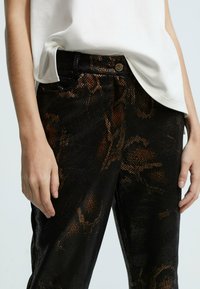 Čierne nohavice s textúrovaným vzorom pripomínajúcim hadiu kožu v odtieňoch zlatej a bronzovej. Majú štandardný pás a predné vrecká, so zapínaním na gombík.