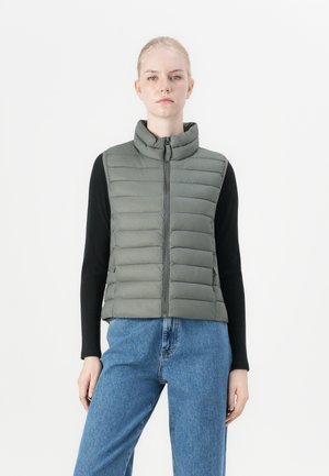 Polstret ermeløs vest i olivengrønn, med høy krage og glidelås foran. Båret over en svart langermet topp, sammen med blå jeans.