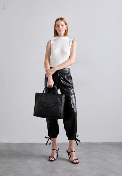 Black leather tote bag with a textured finish, featuring dual handles and a subtle embossed design. Model wears a white top and black pants.