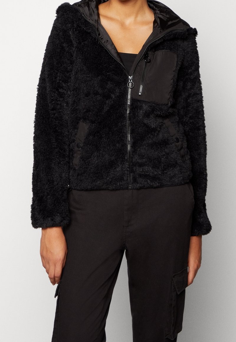 Personne portant une veste noire texturée zippée avec poches latérales et poches poitrine, associée à un pantalon cargo noir, sur un fond uni.