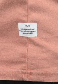 Svijetloružičasta tkanina s mekim teksturom ima ušivenu oznaku na kojoj piše "!SOLID" s proizvodnim detaljima u crnom tekstu.