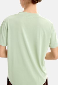 Jasnozielona koszulka z krótkim rękawem, okrągłym dekoltem, gładką fakturą i luźnym krojem, widoczna z tyłu. Brak widocznych wzorów czy dodatków.