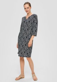 Robe noire avec motif géométrique blanc, décolleté en V, manches trois quarts, longueur genou, tissu léger, associée à des sandales à lanières.