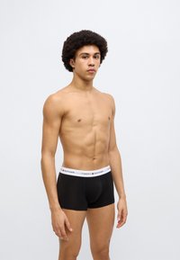 Μαύρα ανδρικά εσώρουχα boxer με λευκή ζώνη μέσης που φέρει το λογότυπο "Tommy Hilfiger". Το μοντέλο έχει μέτριο σώμα και σγουρά μαλλιά. Σχεδίαση σε μονόχρωμο χρώμα.