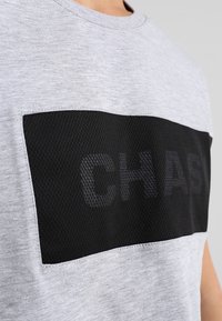 Šedá košile s černou síťovanou obdélníkovou aplikací na hrudi, na které jsou v texturované tkanině částečně viditelné písmena "CHAS".