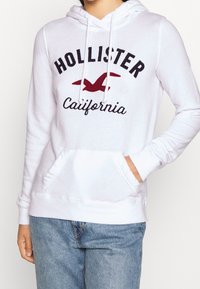 Vit hoodie i mjukt tyg, med framficka, ribbade ärmslut och dragsko i huvan, samt "Hollister California"-grafik på framsidan.