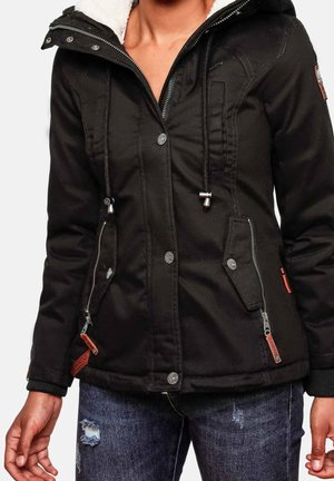 Femme portant une veste à capuche noire avec fermeture éclair frontale, boutons-pression, cordons de serrage et deux poches zippées, associée à un jean bleu déchiré.