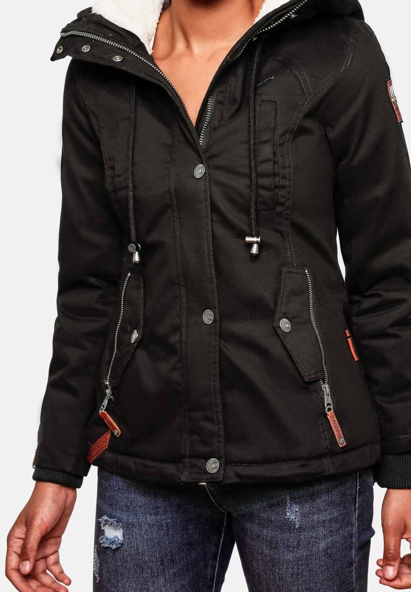Femme portant une veste à capuche noire avec fermeture éclair frontale, boutons-pression, cordons de serrage et deux poches zippées, associée à un jean bleu déchiré.