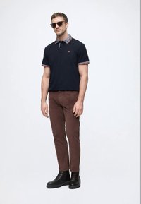 Laivastonsininen poolopaita raidallisella kauluksella, ruskeat corduroy-housut ja mustat saappaat. Henkilöllä on aurinkolasit, minimalistinen muotoilu ja slim fit -leikkaus.