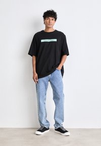 Mustat ylisuuret t-paidat, joissa on mintunvihreä "Calvin Klein" -logo, yhdistettynä vaaleansiniset farkut ja mustat tennarit valkoisilla pohjilla.