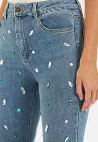 Jeans in denim di colore azzurro, decorati con paillettes di varie forme in tonalità di blu, verde e trasparente, nella tasca frontale e nella zona coscia.