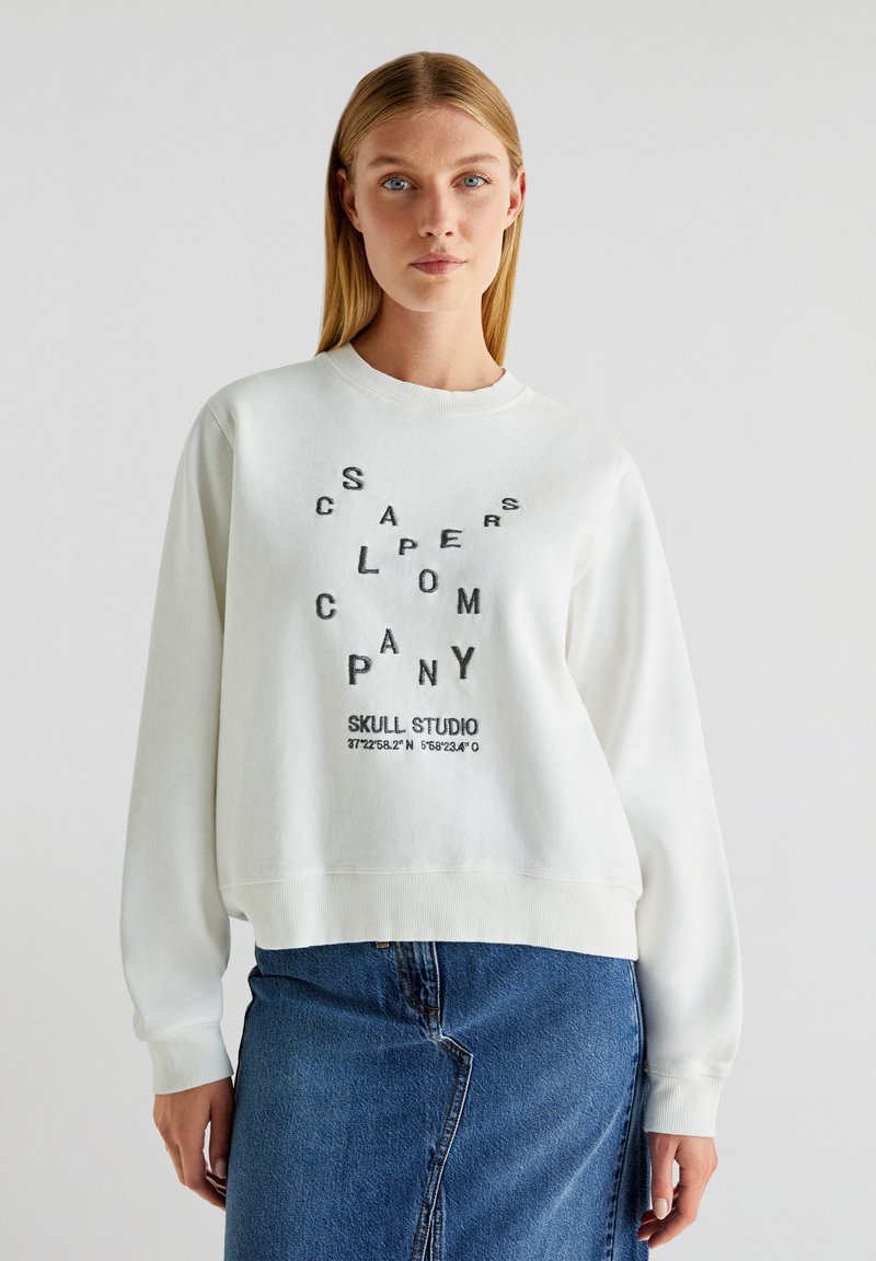 Pulover alb cu text și design grafic negre pe față. Dispune de guler rotund și manșete cu nervuri. Este asortat cu o fustă din denim.