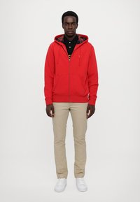 Sudadera roja con cremallera y capucha forrada de cuadros, combinada con una camiseta polo negra, pantalones beige claros y zapatillas blancas. Vista frontal.