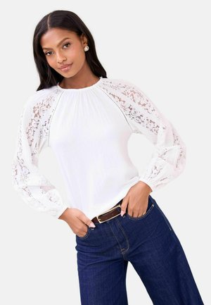 Balta palaidinė su tekstūruotu kūnu ir nėriniais ilgomis rankovėmis. Derinama su tamsiai mėlynais džinsais, išryškinančiais rudos spalvos diržu, neutraliame fone.