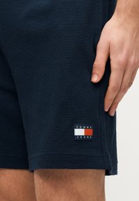 Κοντινό ενός χεριού που ακουμπάει δίπλα σε ναυτικό μπλε υφασμάτινη βερμούδα με σήμα Tommy Jeans κοντά στο στρίφωμα.