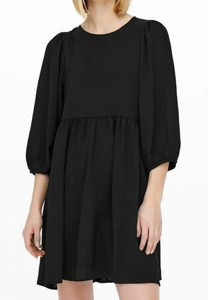 Frau trägt ein locker sitzendes schwarzes Kleid mit rundem Ausschnitt und dreiviertellangen Puffärmeln.