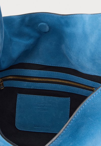 Intérieur d'un sac à main Moschino en daim bleu montrant une poche zippée et une étiquette embossée avec Moschino Milano Made in Italy.