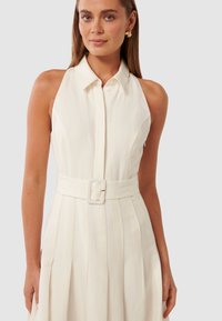 Vestido halter branco com frente abotoada, corpete justo e saia pregueada. Apresenta um cinto largo com fivela retangular. Textura suave.