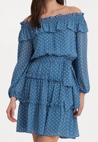 Kvinna klädd i en blå av-axel-klänning med vita prickar, långa ärmar, volangdetaljer och ett knytbälte i midjan.