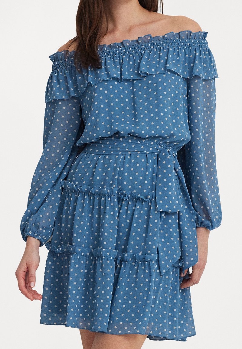 Kvinna klädd i en blå av-axel-klänning med vita prickar, långa ärmar, volangdetaljer och ett knytbälte i midjan.