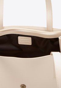 Borsa a spalla in pelle beige con due manici, che mostra un interno marrone con un'etichetta del marchio. Dispone di una chiusura a scatto e bordi cuciti.