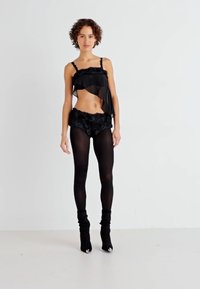 Camiseta negra transparente con acentos de encaje, combinada con pantalones cortos de patrón floral. La modelo lleva medias negras y botines de tacón alto. Fondo blanco liso.