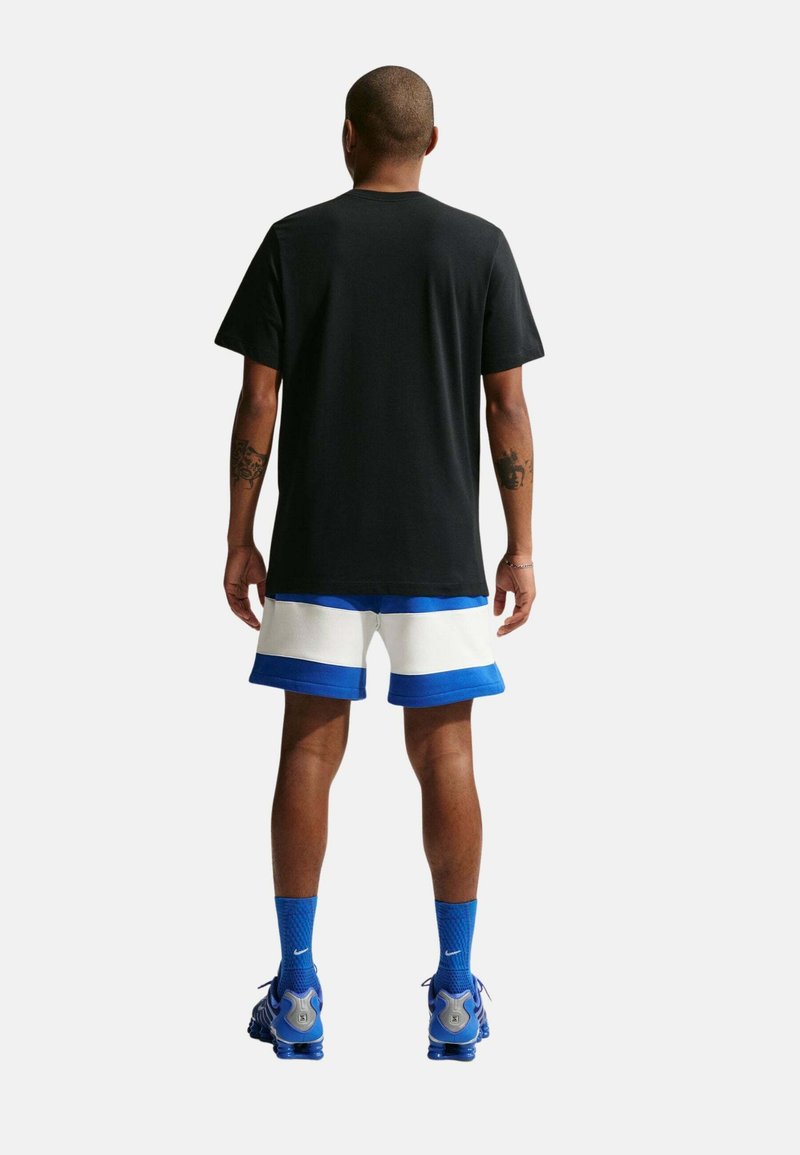 Hombre de pie de espaldas vistiendo una camiseta negra, pantalones cortos blancos y azules, calcetines azules de Nike y zapatos deportivos azules sobre fondo blanco.