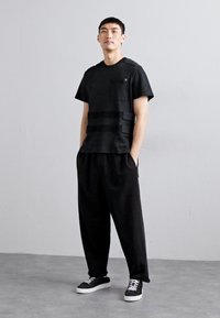 Schwarzes T-Shirt mit kurzen Ärmeln und strukturierten Akzenten sowie horizontalen Streifen, kombiniert mit lockeren schwarzen Hosen und schwarzen Sneakern mit weißen Sohlen.