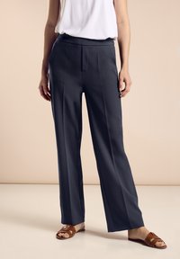 Vidbenade byxor i mörkgrått tyg med en slät yta. Har en rak front och vertikala sömmar. Matchas med bruna sandaler.