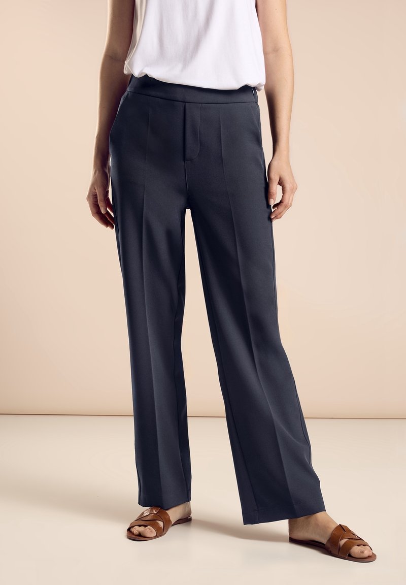 Vidbenade byxor i mörkgrått tyg med en slät yta. Har en rak front och vertikala sömmar. Matchas med bruna sandaler.