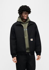 Μαύρο μπουφάν με φερμουάρ και υφή, με μεγάλη μπροστινή τσέπη και λογότυπο. Φοριέται πάνω από μια ελιά πράσινη κουκούλα.