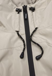 Hellbeige Jacke mit einem Front-Reißverschluss, ausgestattet mit schwarzen Akzenten, verstellbaren Zugbändern mit schwarzen Stoppern und glattem, wasserabweisendem Stoff.