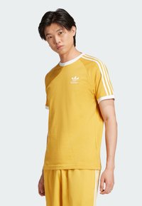 Keltainen puuvillapaita, jossa on valkoinen reunus ja kolme valkoista raitaa hihoissa. Paidassa on pieni valkoinen Adidas-logo rinnassa.