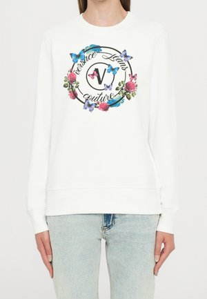 Biała bluza z długim rękawem z kolorowym kwiatowym i motylim wzorem otaczającym tekst "Versace Jeans Couture" z przodu, noszona z jasnoniebieskimi jeansami.