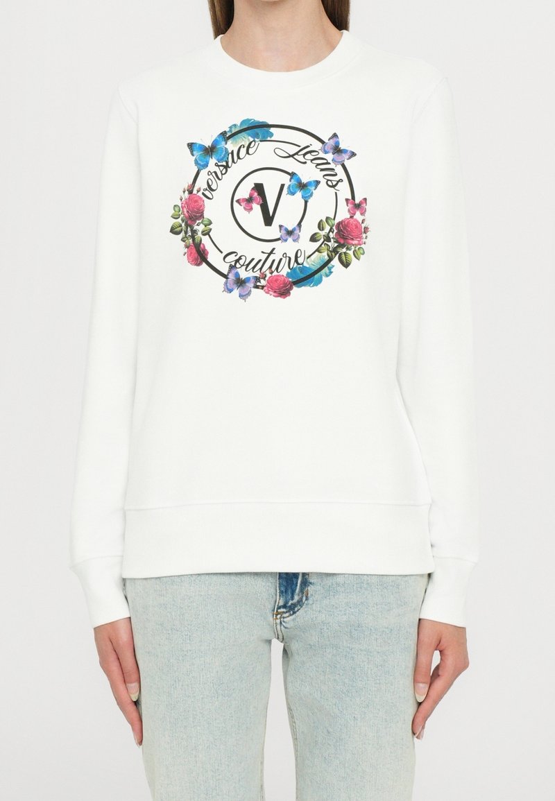 Sweat-shirt blanc à manches longues avec un motif floral et papillon coloré entourant le texte "Versace Jeans Couture" à l'avant, porté avec un jean bleu clair.