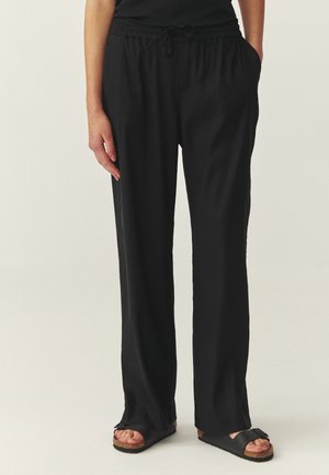 Osoba v nesúdržných čiernych nohaviciach so šnúrkou a čiernych obuviach slip-on s korkovými podrážkami stojaca na svetlom pozadí.