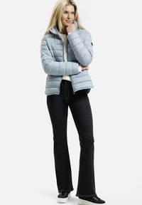 Blondynka w jasnoniebieskiej pikowanej kurtce, czarnych rozszerzanych dżinsach i czarnych trampkach, stojąca z założonymi rękami i spoglądająca na bok.