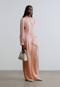 Ferskenfarvet satinbluse med mandarin-krave, kombineret med viddebukser i samme farve. Modellen holder en lille hvid håndtaske for kontrast.