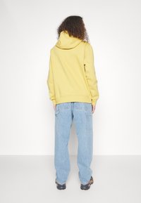 Sudadera amarilla con capucha de ajuste relajado, combinada con vaqueros azul claro de corte ancho. La textura parece suave y casual.