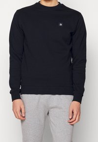 Sudadera negra de algodón, con mangas largas, cuello y dobladillo acanalados, y un pequeño parche con logo en el pecho. Combinada con pantalones grises.