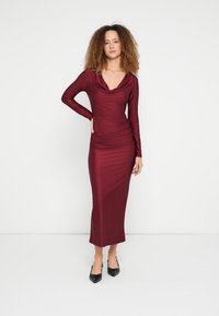 Μακρυμάνικο μπορντό φόρεμα με λαιμόκοψη κασκόλ, εφαρμοστή γραμμή και ρυτιδωμένη πλευρική λεπτομέρεια, συνδυασμένο με μαύρα επίπεδα παπούτσια. Ομαλή υφή υφάσματος.