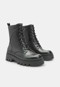 Un paio di stivali da combattimento neri in pelle con lacci, suole spesse e cerniere laterali su uno sfondo neutro.