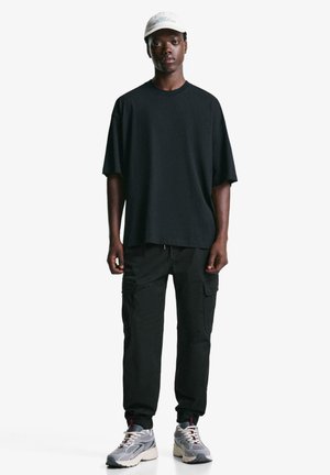 Tricou oversize negru, pantaloni cargo negri cu buzunare laterale, adidași gri cu accente roșii și o șapcă de culoare deschisă.