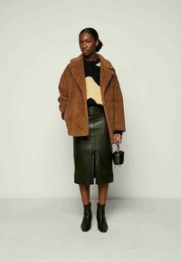 Kvinne iført en brun pelsjakke i kunstig pels, svart og beige mønstret genser, mørkegrønn lærskjørt, svarte ankelstøvler, holder en liten svart håndveske.