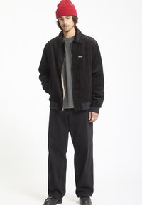 Zwarte corduroyjas met een ritssluiting, grijs shirt eronder, zwarte wide-leg broek en een rode gebreide muts. Minimalistische ontwerpelementen.