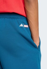 Teal sportieve shorts met een ritszak en witte adidas-logo. Marvel-merkgeving in het rood op de linker bovenbeen. Zacht materiaal en elastische tailleband.