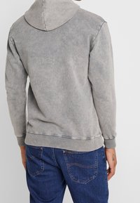 Grijze capuchontrui met een zachte textuur, geribbelde manchetten en zoom, met een minimalistisch ontwerp en verfrommeld uiterlijk. Draagt blauw spijkerbroek.